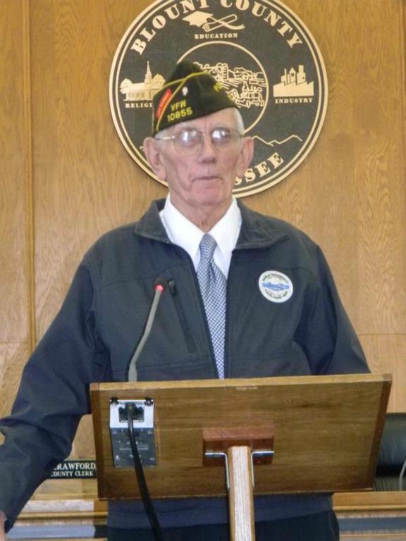 Man in blue jacket and white shirt, with glasses and hat, stands speaking at podium